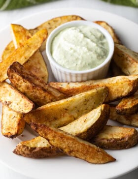 A white plate piled with thick-cut air fryer potato wedges, with a white ramekin of white dipping sauce also on the plate.