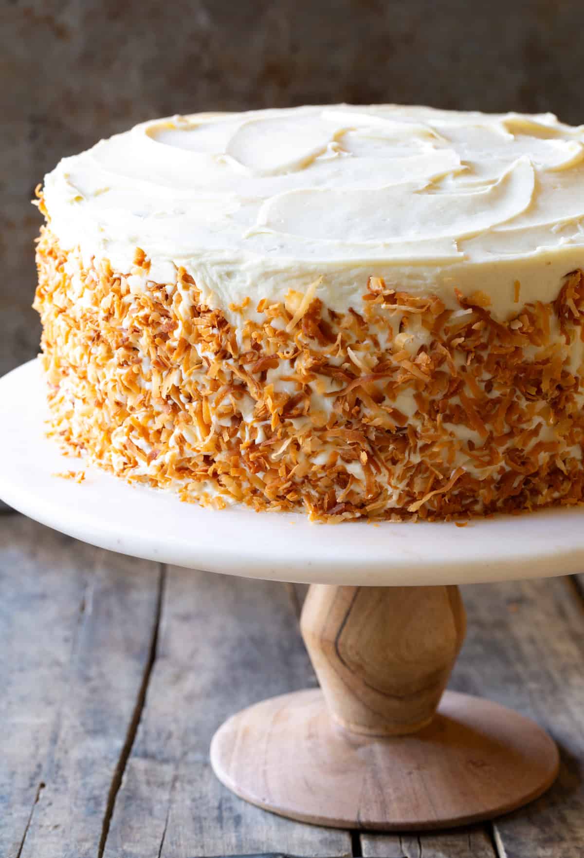 Three layer easy carrot cake recipe covered in white frosting and toasted coconut. The cake is on a white pedestal cake stand.