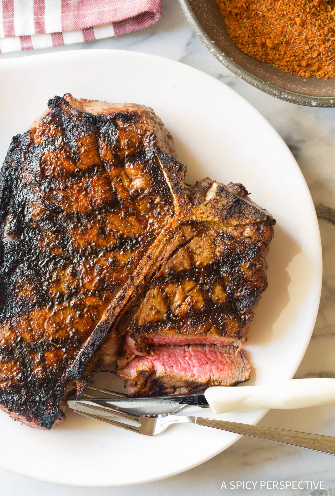 Steak #ASpicyPerspective #Steak #SteakSeasoning #HowtoSeasonSteak #BestSteakSeasoning #SteakSeasoningRecipe #HomemadeSeasoning #Grilling #GoodSteakSeasoning Overhead shot of steak on a white plate with fork and knife.