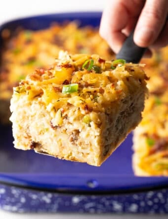 A slice of hashbrown breakfast casserole is being held by a metal spatula above a blue baking dish.