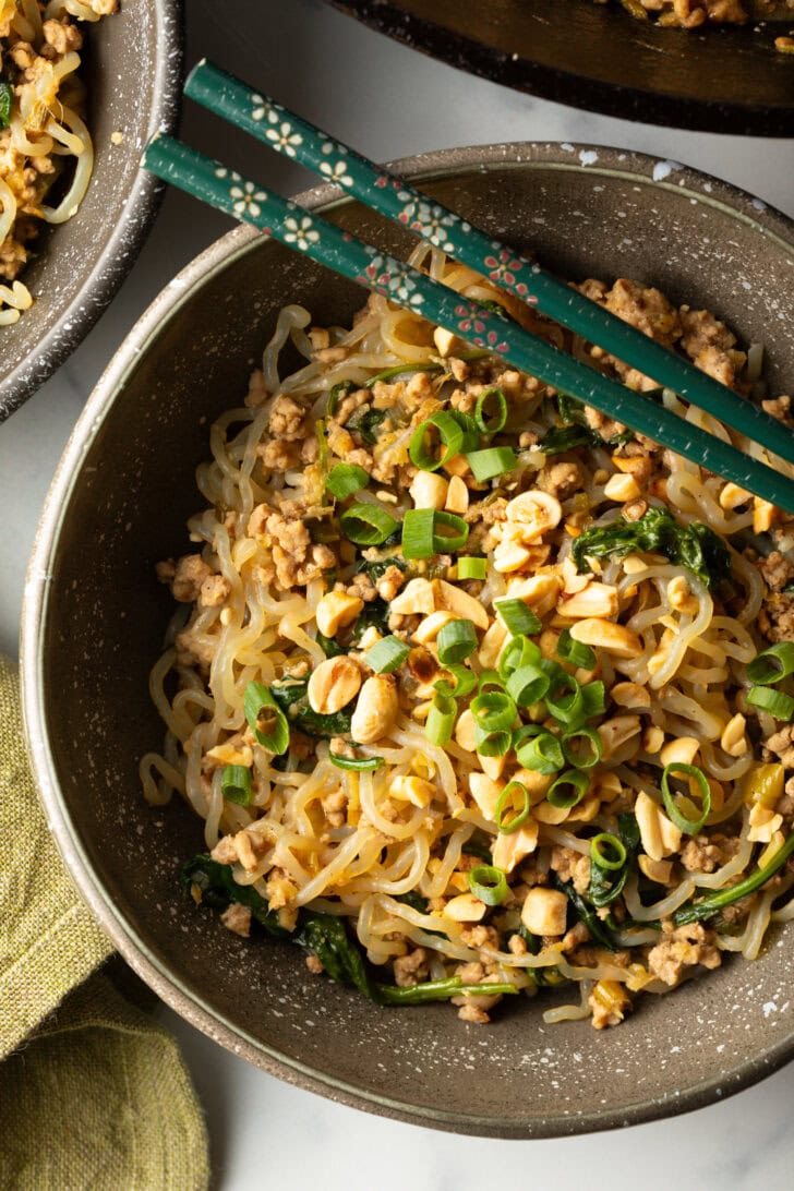 Spicy Szechuan noodles in a black bowl topped with green onion slices. Set of chopsticks are resting on top of the bowl.