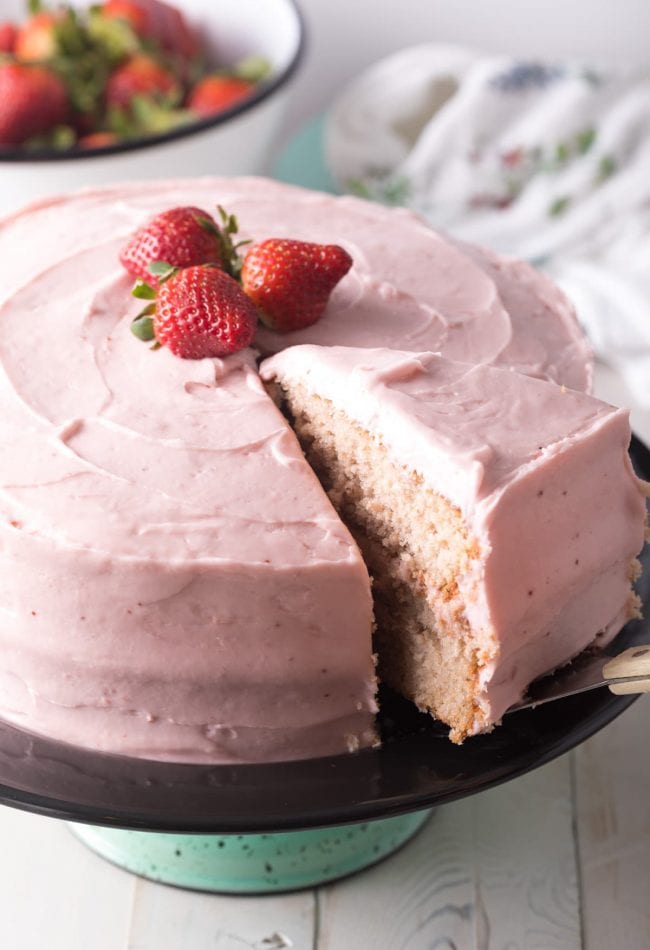 Whole fresh strawberry cake with pink frosting, topped with three fresh strawberries. A slice has been cut and is being removed by a cake slicer.