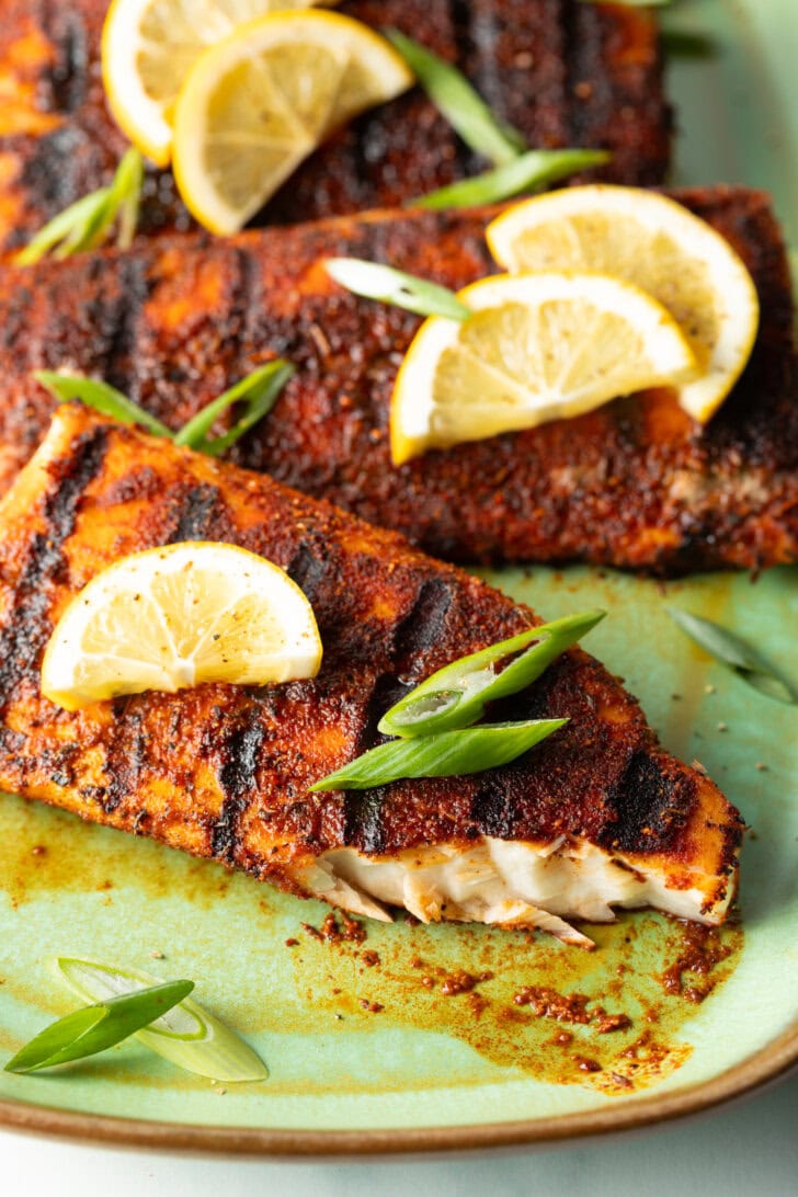 Close view of a fillet of grilled mahi mahi topped with a slice of lemon and green onions. A bite has been removed from the fish to show the white flaky inside of the meat.