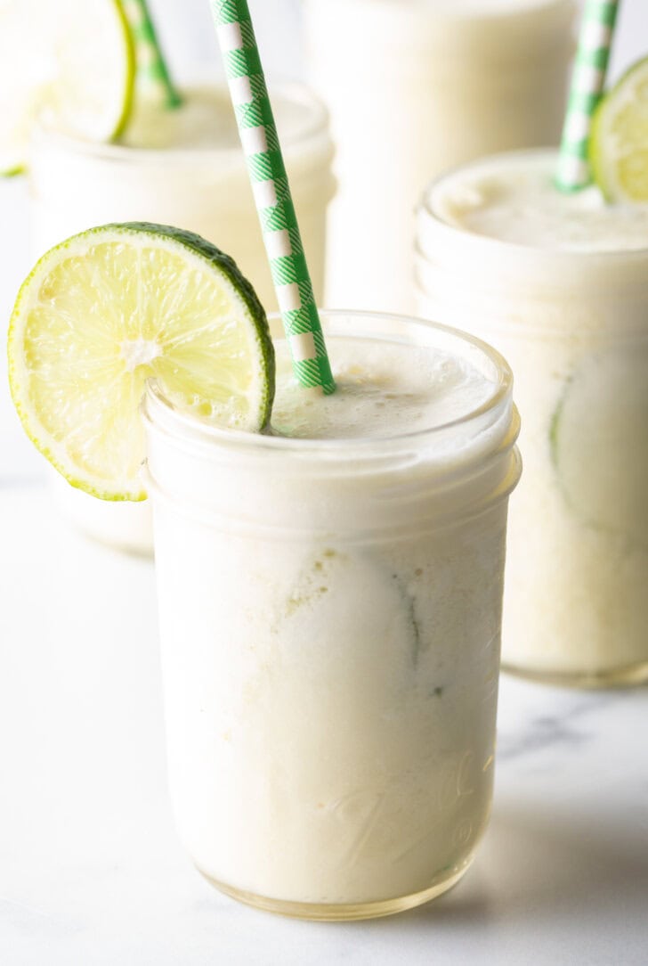 Four glasses of Brazilian limeade, each garnished with a slice of lime and a green and white striped straw.