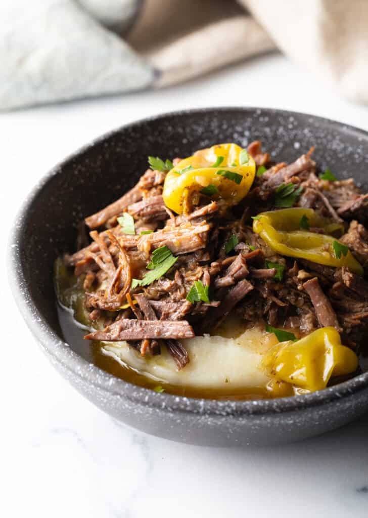Black bowl with a layer of mashed potatoes topped with shredded pot roast and pepperoncinis.