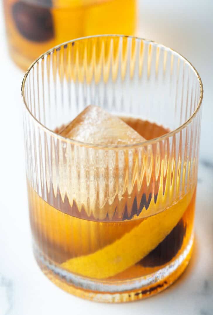 Old fashioned cocktail in a lowball glass, with single large cube of ice, cherry and orange peel in the glass.