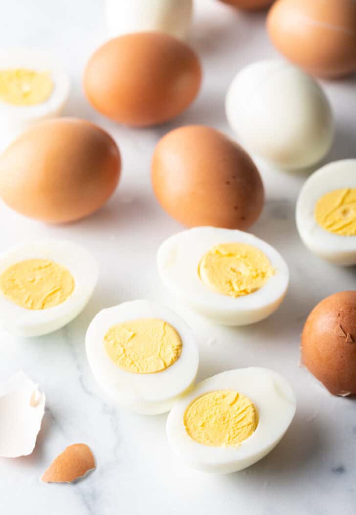 A dozen hard boiled eggs on a white marble backdrop. There are five egg halves, four brown eggs in the shell, and one white peeled egg.